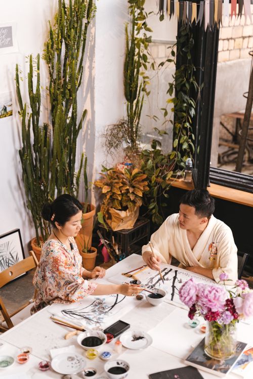 Free Two artists collaborating in a brightly lit, plant-filled art studio, sharing ideas and painting together. Stock Photo