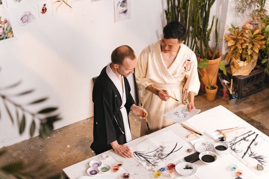 Two artists in robes collaborate on a painting in a bright, plant-filled studio.