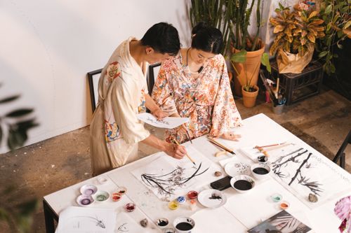 Free Two Asian artists in robes collaborating on a painting in an art studio using various brushes and paints. Stock Photo