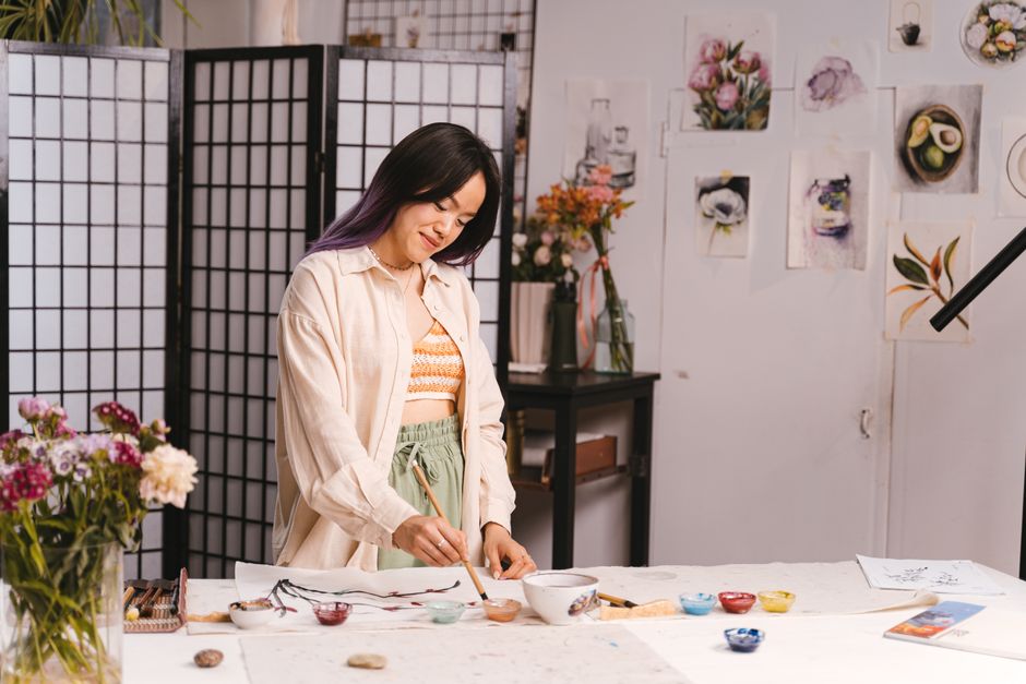 An Asian female artist painting at a work table in a vibrant art studio.