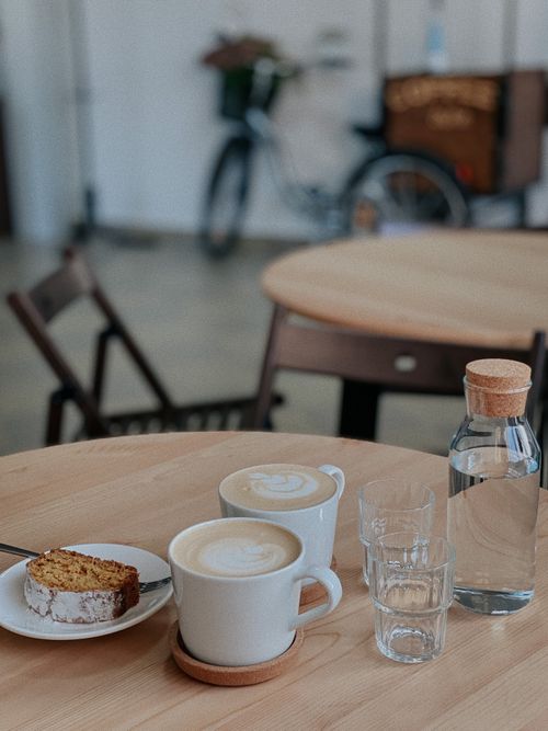 Free Warm café scene featuring lattes, cake slice, and glass water bottle on a wooden table. Stock Photo