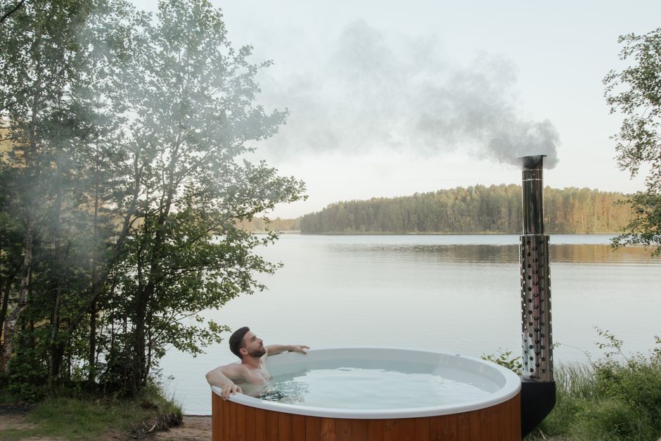 Man enjoying a serene soak in an outdoor hot tub by a peaceful lake surrounded by nature.
