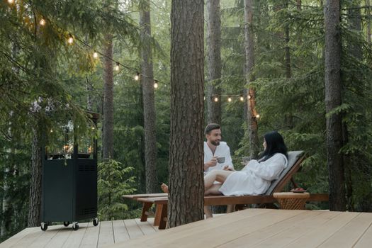 Couple in bathrobes enjoying relaxation on lounge chairs in serene forest setting.