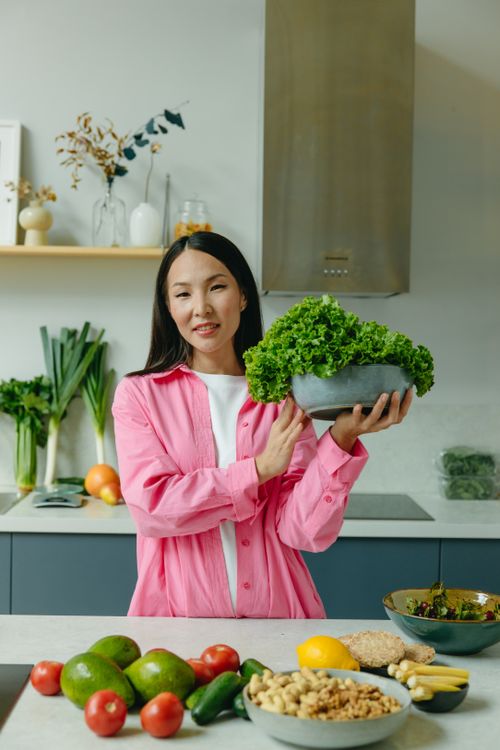 Free Asian woman in pink sleeves holding lettuce in a kitchen filled with fresh organic vegetables. Stock Photo