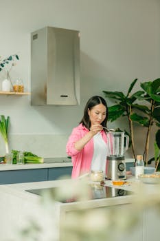 Asian woman in pink overshirt blending a healthy smoothie in a modern kitchen setting.