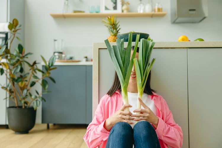 Smiling Woman Sitting On The Floor Covering Face With Leeks