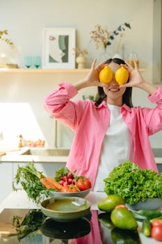 A cheerful woman playfully holds lemons over her eyes in a bright kitchen with fresh vegetables.
