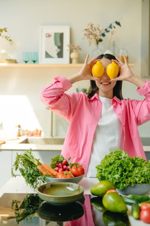 Free A cheerful woman playfully holds lemons over her eyes in a bright kitchen with fresh vegetables. Stock Photo