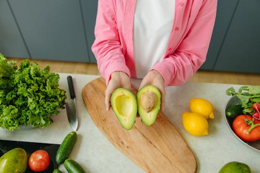 Person holding a sliced avocado on a kitchen counter with fresh vegetables around.