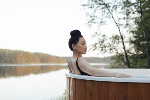 Asian woman enjoying a serene moment in a hot tub overlooking a tranquil lake.