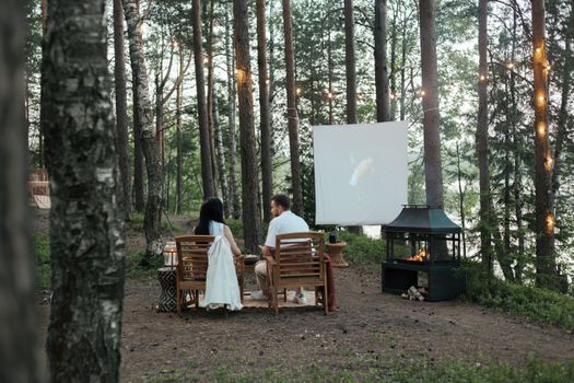 Couple enjoying a cozy movie night in a forest setting with a warm fireplace.