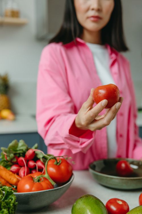 Free A person holding a fresh tomato over a bowl of assorted vegetables in a kitchen setting. Stock Photo