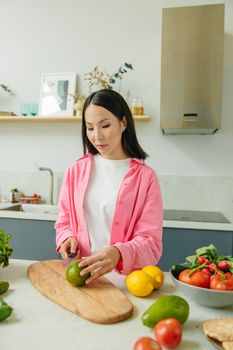 Asian woman in pink overshirt slicing vegetables in a bright kitchen.