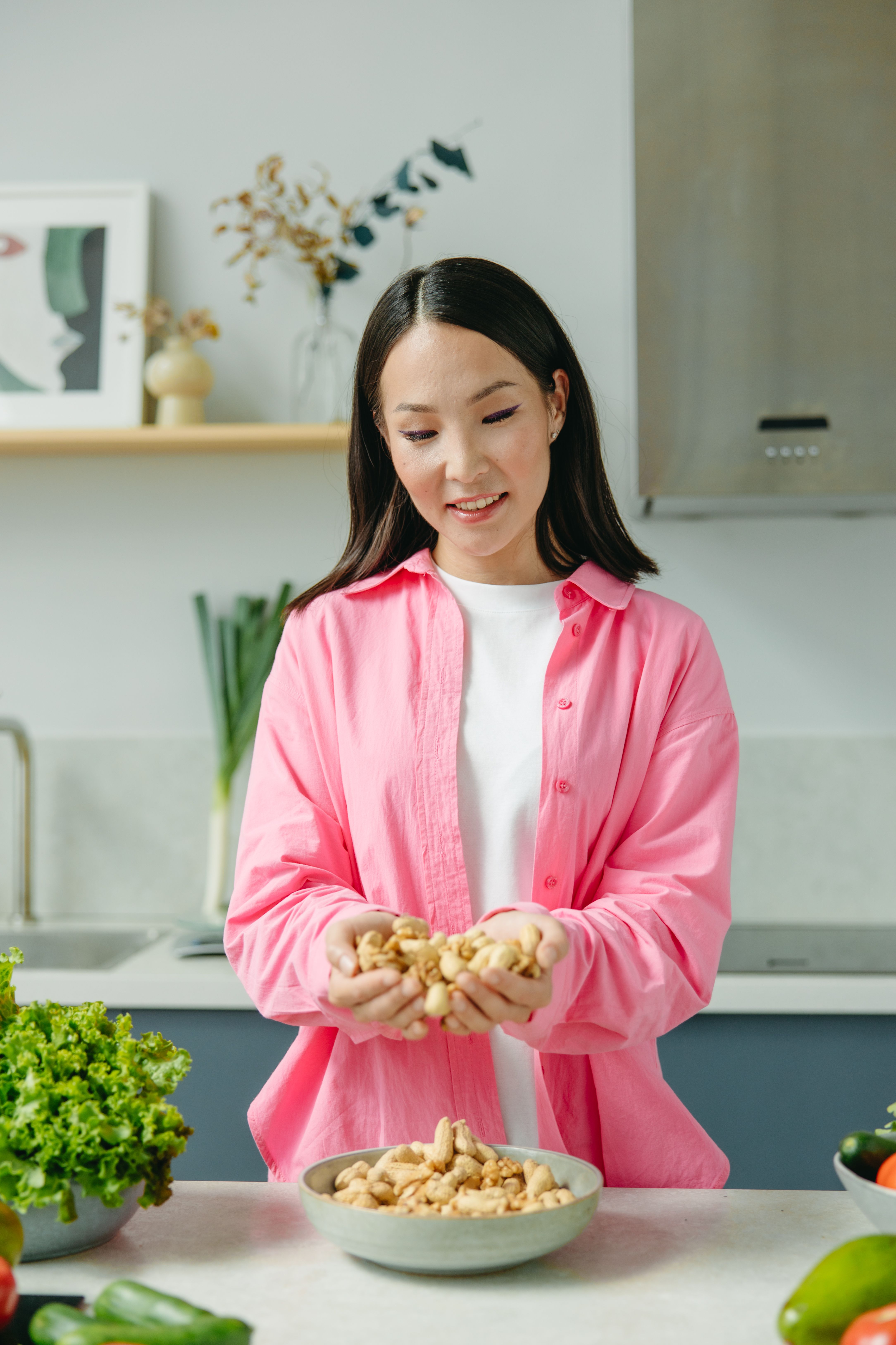 Young Asian woman in kitchen holding a bowl of peanuts and enjoying fresh vegetables.