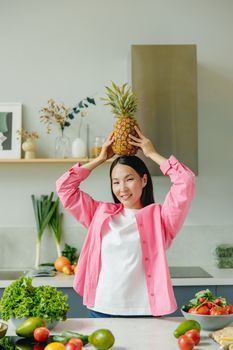 Asian woman playfully holding a pineapple on her head amid fresh produce in the kitchen.