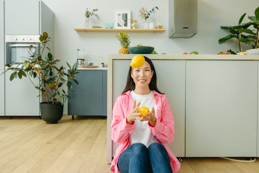 Smiling woman juggles lemons in a modern kitchen with plants and wooden flooring.