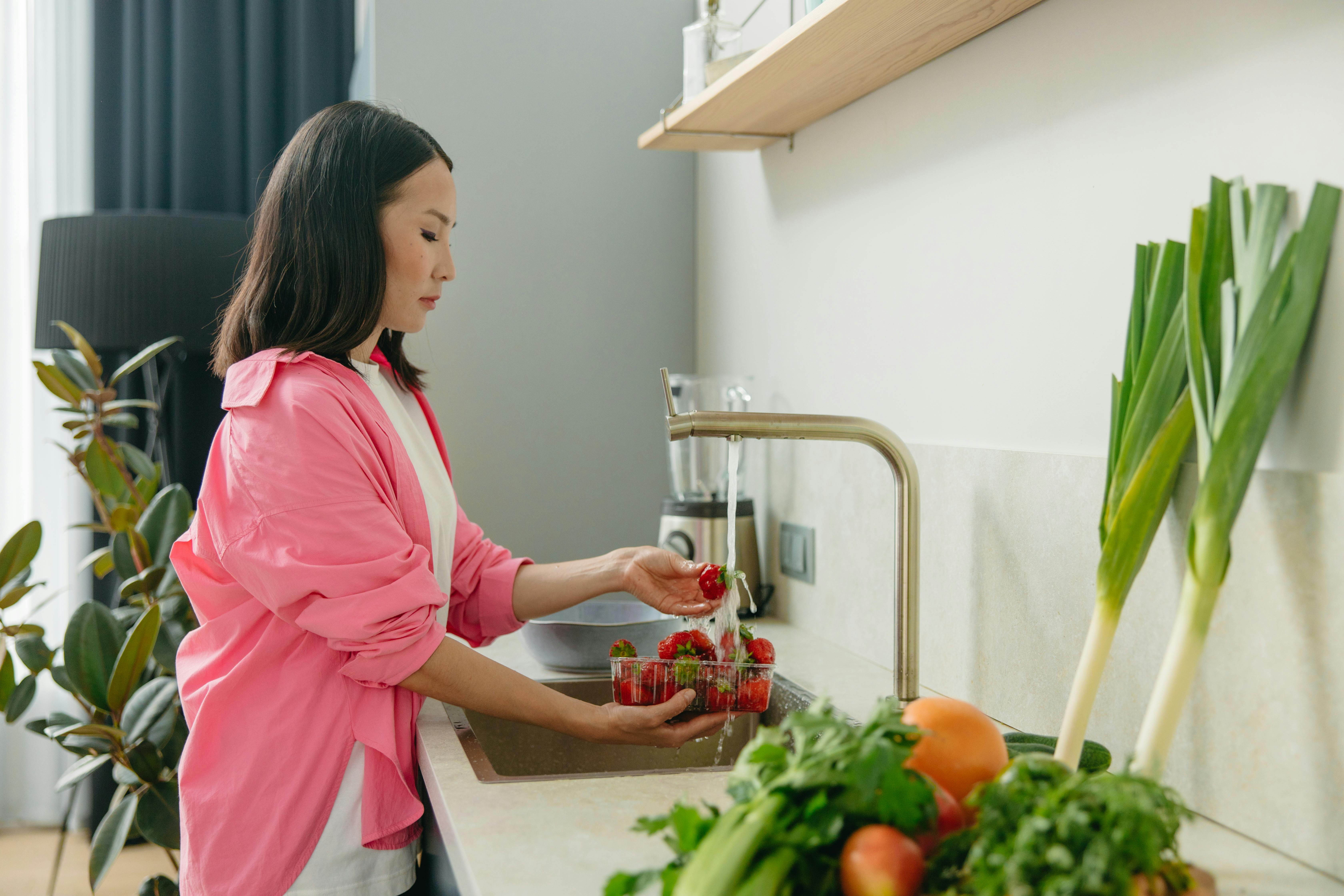 Asian woman washes strawberries at kitchen sink, promoting healthy living.