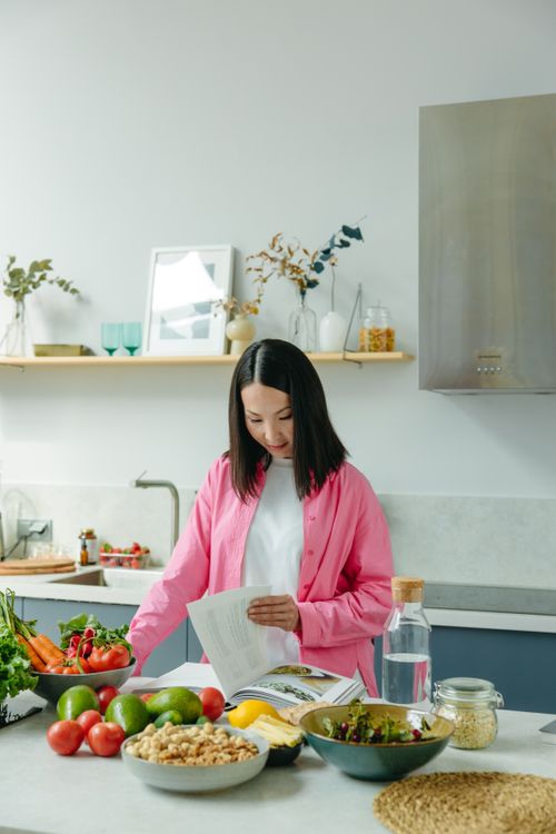 Free Woman in pink shirt preparing fresh vegetables and reading a recipe in a modern kitchen. Stock Photo