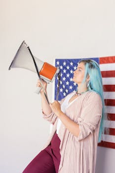 Caucasian woman with blue hair holding a megaphone in front of an American flag indoors.