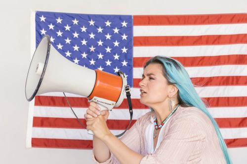 Free Expressive woman using a megaphone in front of a vibrant American flag, symbolizing freedom. Stock Photo