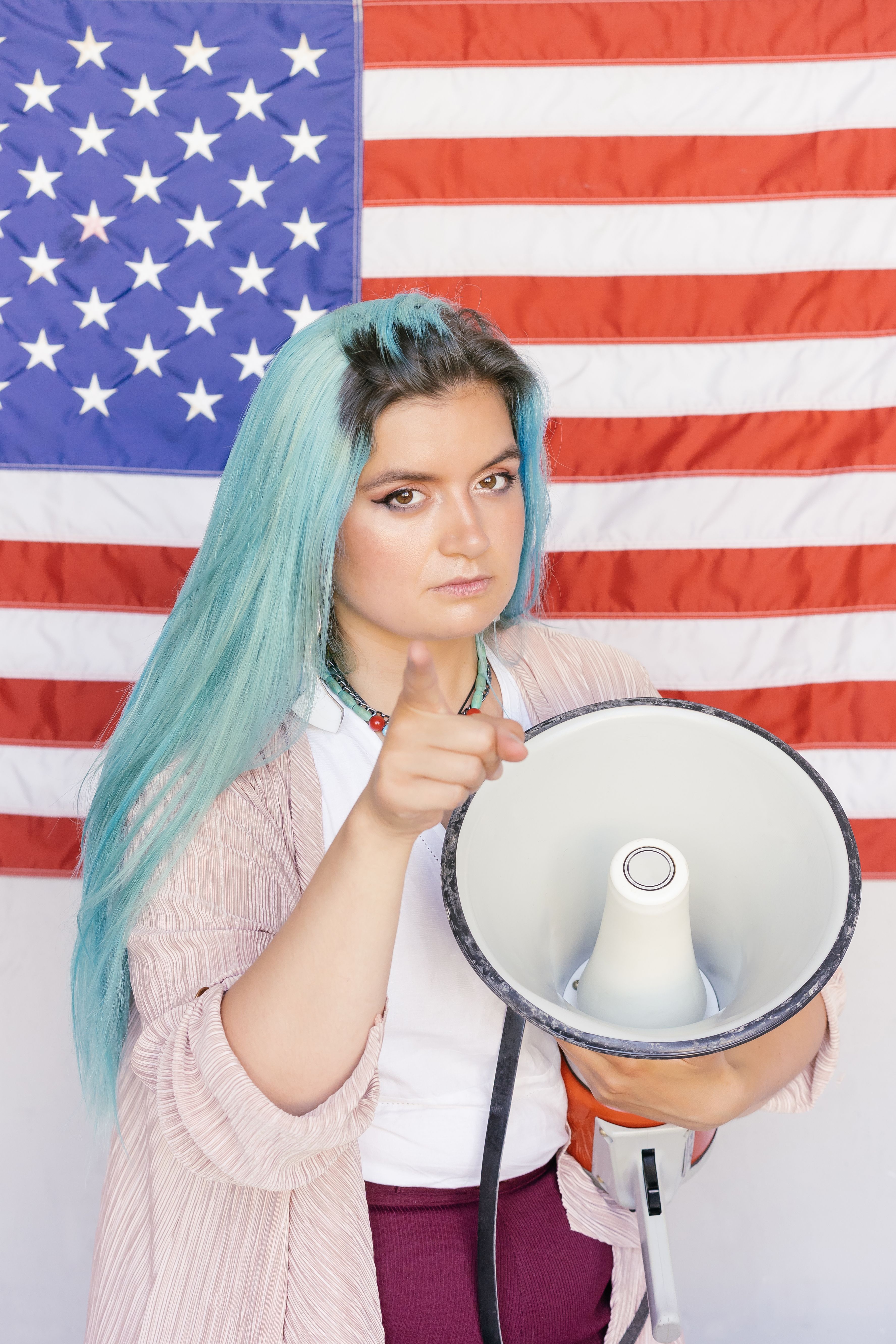 Caucasian woman with blue hair holds megaphone and points, standing in front of an American flag.