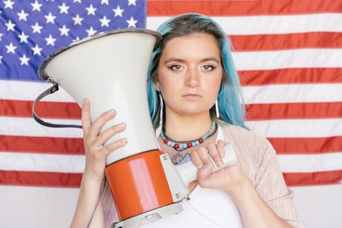 Free Caucasian woman holding a megaphone in front of an American flag, symbolizing freedom of speech. Stock Photo
