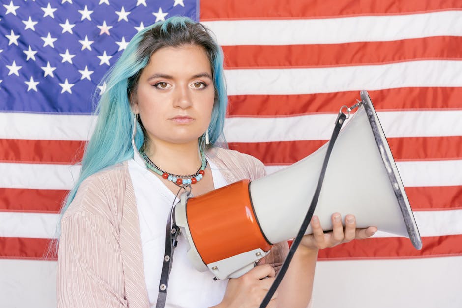 Caucasian woman with blue hair holding a megaphone in front of an American flag, symbolizing activism.