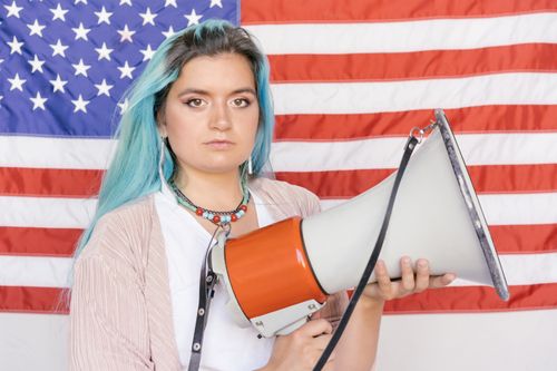 Free Caucasian woman with blue hair holding a megaphone in front of an American flag, symbolizing activism. Stock Photo