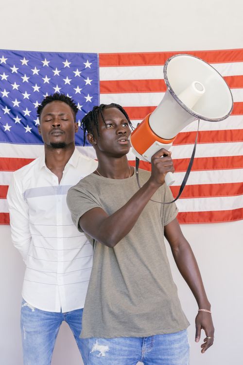 Free Two young men stand with a megaphone in front of an American flag, exuding confidence and activism. Stock Photo