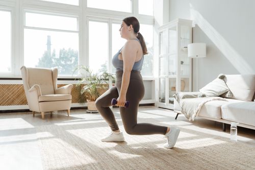 Free A woman in activewear doing lunges with dumbbells in a bright living room. Stock Photo