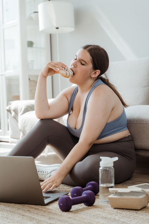 Free A woman in activewear enjoying a snack during a workout break at home, surrounded by dumbbells and a laptop. Stock Photo