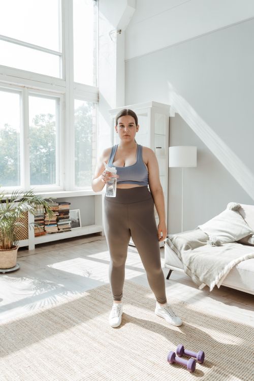 Free Confident woman in activewear holding a water bottle, standing in a sunlit living room with dumbbells. Stock Photo