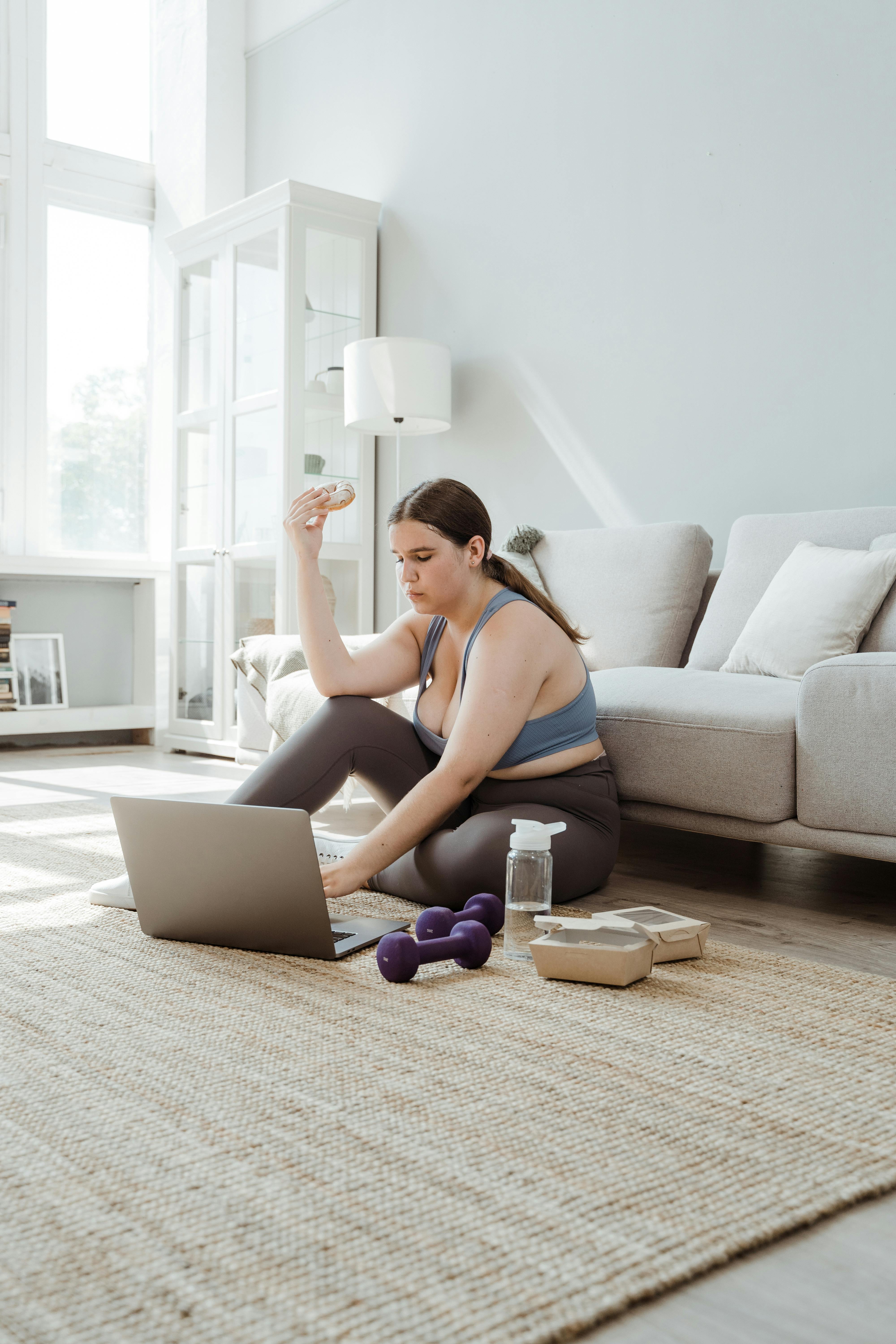 woman in sportswear sitting on floor using laptop and eating doughnut