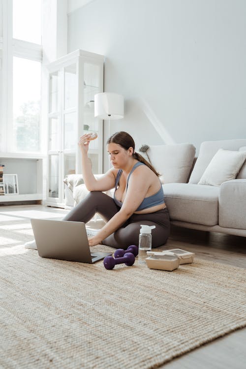 Free Woman in Sportswear Sitting on Floor Using Laptop and Eating Doughnut Stock Photo