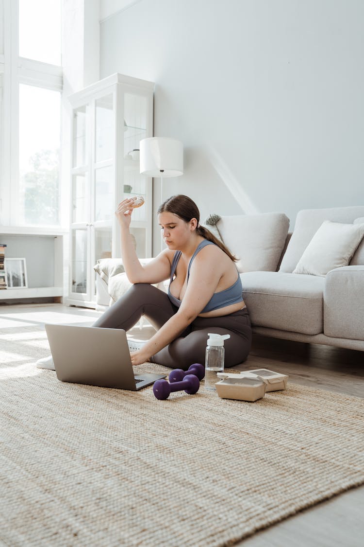 Woman In Sportswear Sitting On Floor Using Laptop And Eating Doughnut