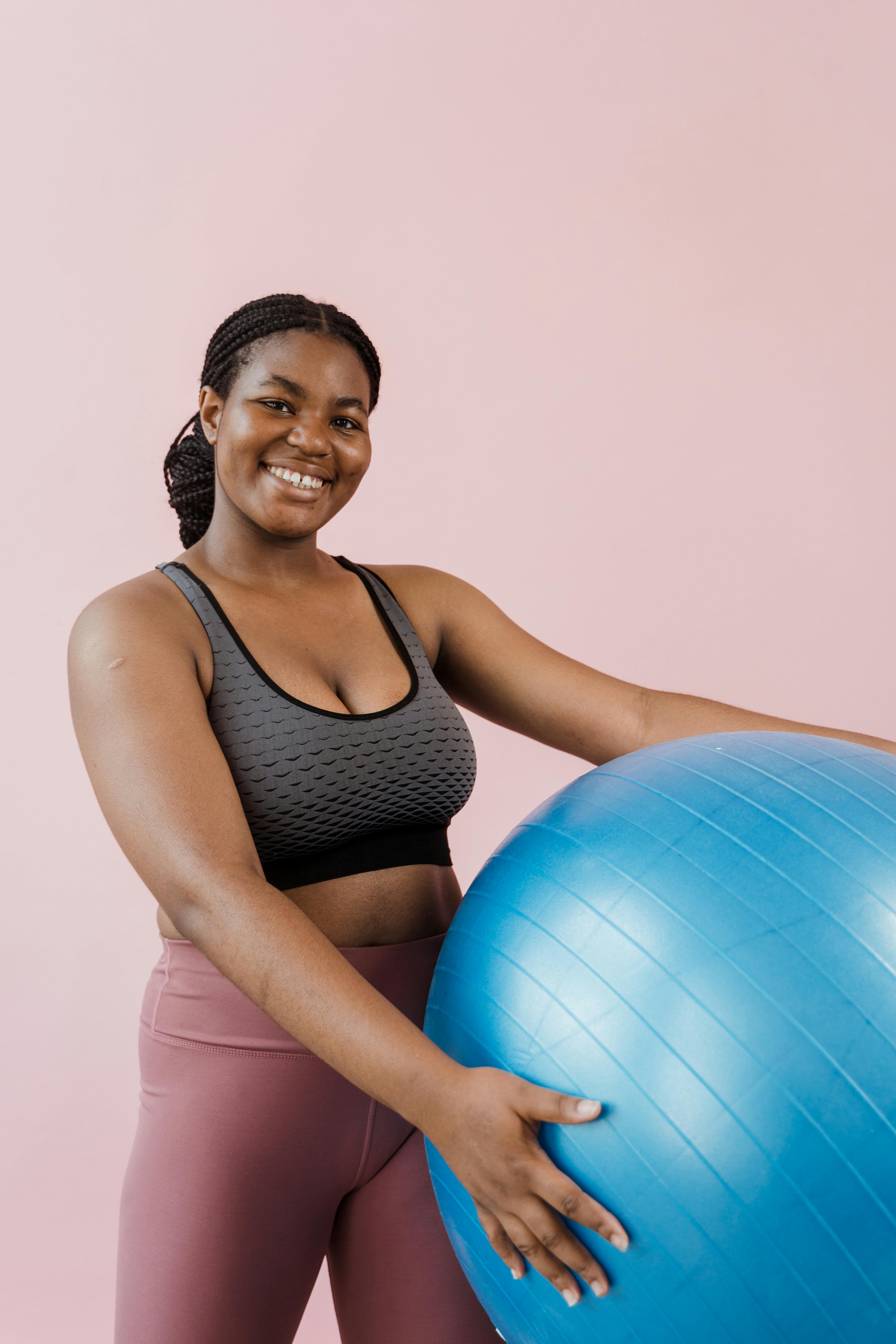 一位面带微笑的非裔美国女性，在淡粉色的背景下拿着一个蓝色的健身球，穿着运动服