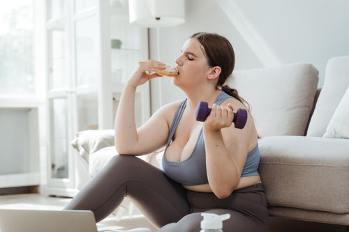 Free Confident plus-size woman enjoying donut while exercising at home. Stock Photo