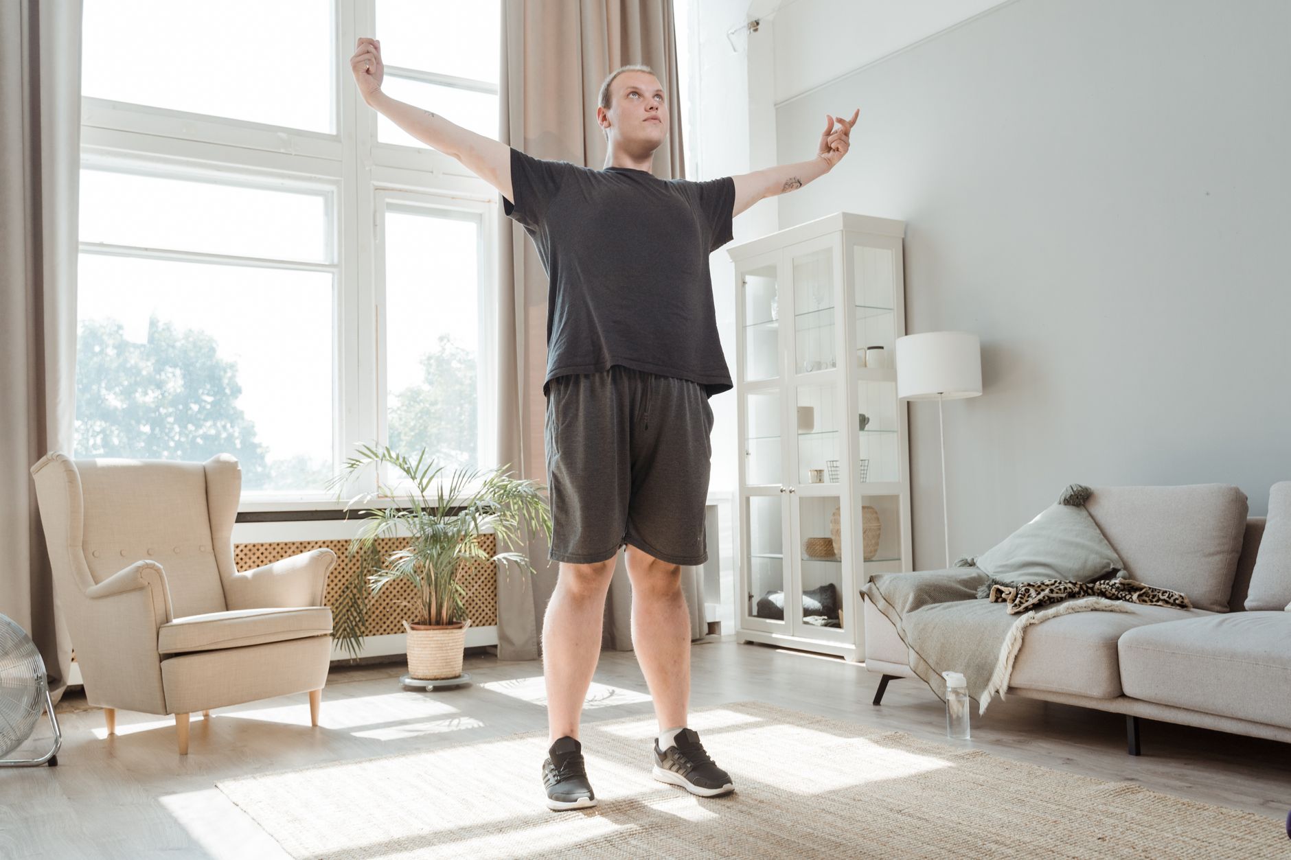 A man performs a stretching exercise with arms out in a bright, modern living room.