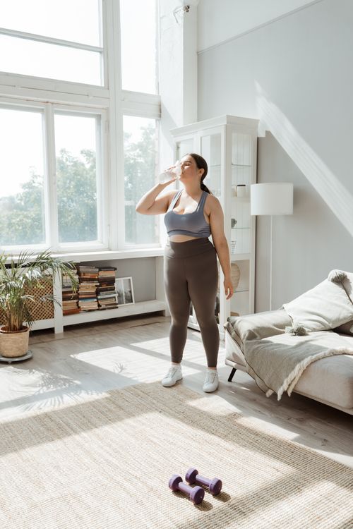 Free Woman in activewear rehydrating post-workout in a sunny living room setting. Stock Photo