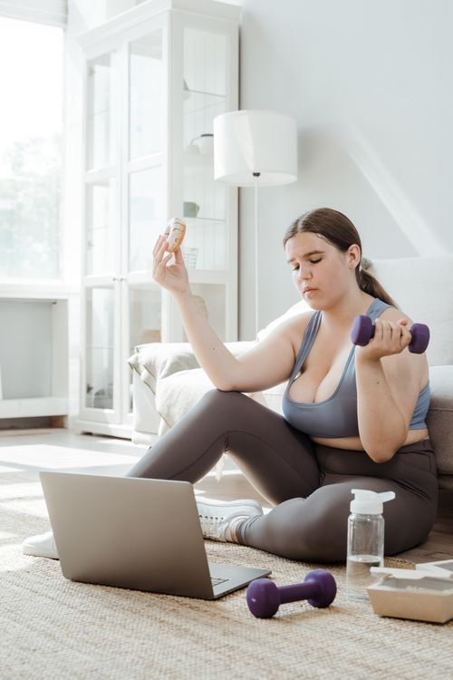 Free A plus size woman at home balancing fitness with lifestyle, using weights and enjoying a donut. Stock Photo