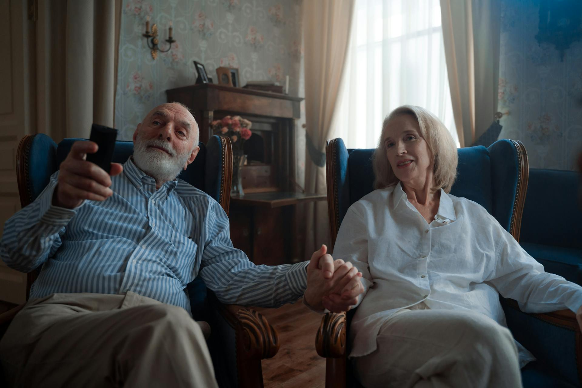 Senior couple sitting together, holding hands and watching TV in a cozy living room.