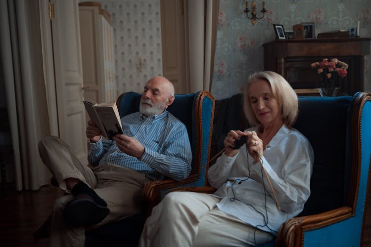 An Elderly Couple Sitting On The Chair While Knitting And Reading A Book