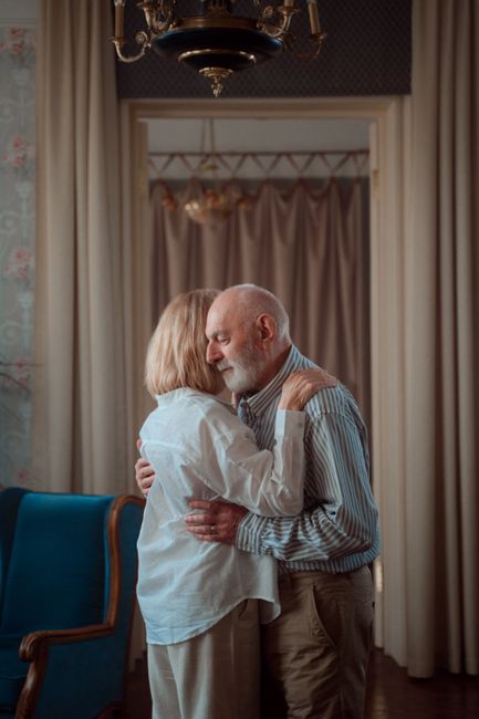 An older couple embracing in a comfortable, elegantly furnished living room.