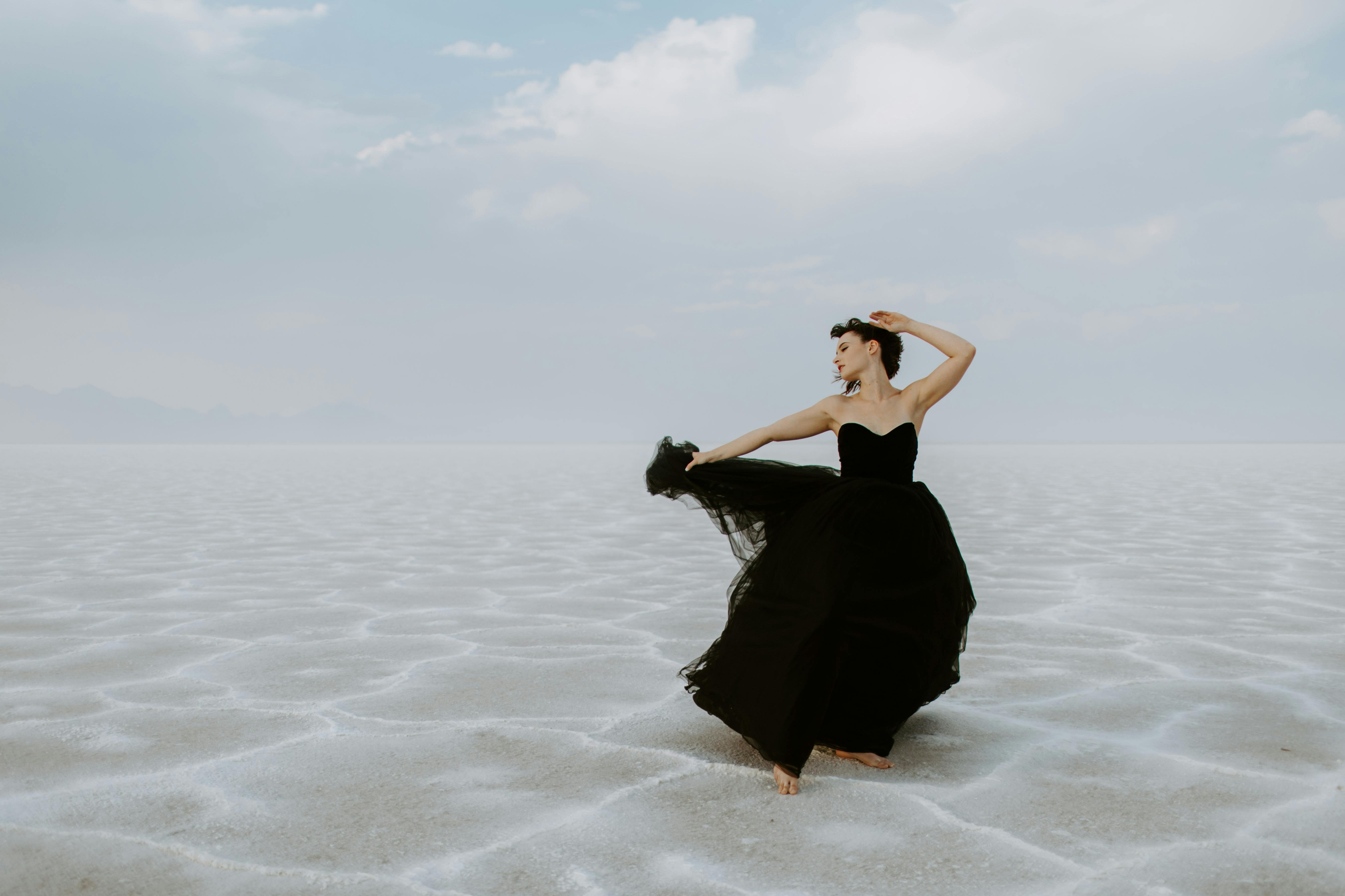 A woman in a black gown poses gracefully on vast salt flats under a cloudy sky, creating a striking outdoor scene.