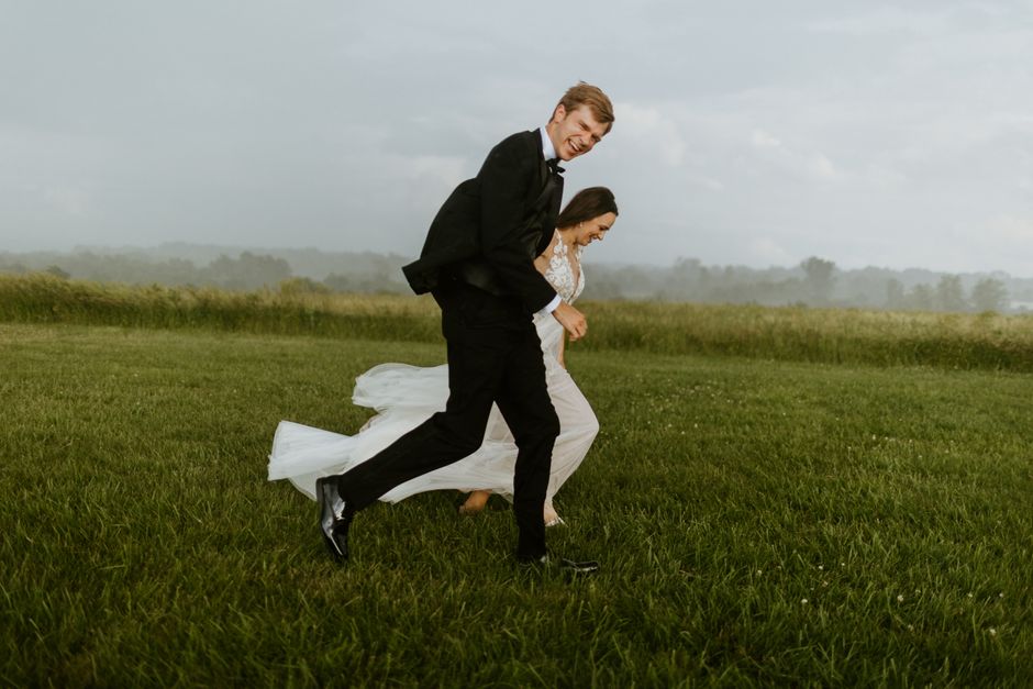 A joyful couple runs through a grassy field on their wedding day, capturing a candid moment.