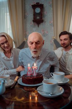 Senior man blowing candles on birthday cake with family indoors, celebrating togetherness.