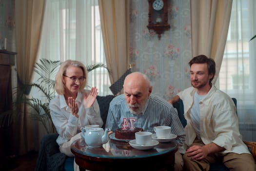 Elderly man blows out candles on cake surrounded by family indoors.