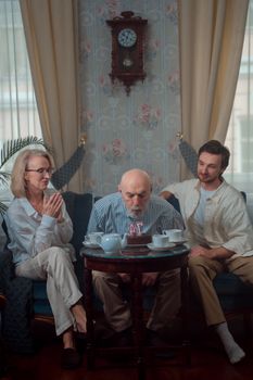 A senior man blows out candles on a birthday cake with family indoors.