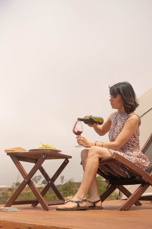 A Woman in Floral Dress Sitting on a Wooden Chair while Pouring a Wine on a Glass