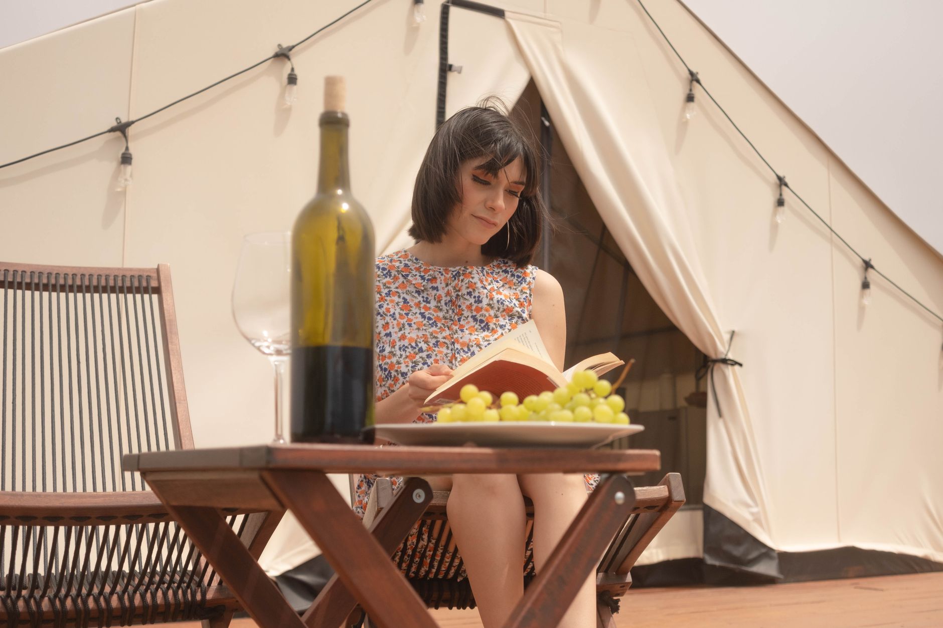 A woman enjoys a book with wine and grapes at an outdoor campsite, relaxing in a floral dress.